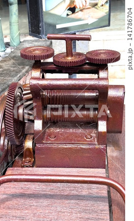 Vintage metal gear for producing silver mechanism with threaded rollers, bevel gears, toothed handwheels, cast frame, and wooden base, showing rusty industrial engineering details from top front view Vintage metal gear for producing silver mechanism with threaded rollers, bevel gears, toothed handwheels, cast frame, and wooden base, showing rusty industrial engineering details from top front view 141786704