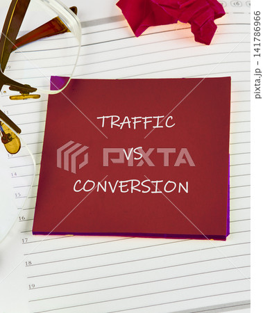 A red sticky note shows the words traffic vs conversion on a white notebook. Glasses and a pink flower are nearby. This scene is in an office during the day 141786966