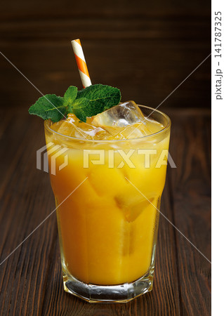 Orange juice in glass with ice cubes, fresh mint sprigs and striped straw on dark wooden table Orange juice in glass with ice cubes, fresh mint sprigs and striped straw on dark wooden table 141787325