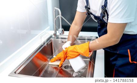 Male housekeeper wearing orange rubber gloves holding spray bottle while scrubbing on kitchen sink Male housekeeper wearing orange rubber gloves holding spray bottle while scrubbing on kitchen sink 141788158