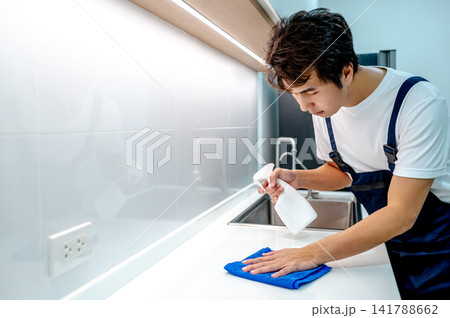 Asian male housekeeper holding spray bottle while wiping microfiber cloth on white countertop Asian male housekeeper holding spray bottle while wiping microfiber cloth on white countertop 141788662