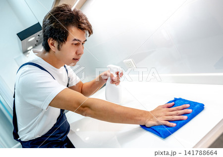 Asian male housekeeper holding spray bottle while wiping microfiber cloth on white countertop 141788664