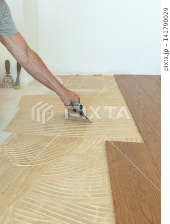 Worker applying adhesive on concrete floor with notched trowel during oakwood parquet installation Worker applying adhesive on concrete floor with notched trowel during oakwood parquet installation 141790029