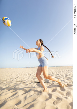 Young woman plays volleyball on the beach during a sunny day while people walk in the background near the water Young woman plays volleyball on the beach during a sunny day while people walk in the background near the water 141790076
