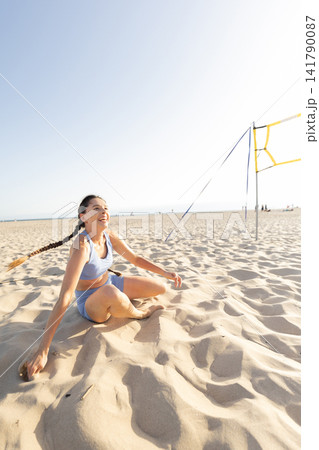 Person enjoys time on the beach while sitting in soft sand near a volleyball net under a clear blue sky Person enjoys time on the beach while sitting in soft sand near a volleyball net under a clear blue sky 141790087