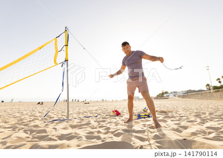 Person sets up a beach volleyball net while others relax on the sand on a sunny day Person sets up a beach volleyball net while others relax on the sand on a sunny day 141790114