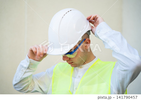 Worker adjusting hard hat while preparing for work in safety gear. Personal protective equipment and safety training in a construction environment Worker adjusting hard hat while preparing for work in safety gear. Personal protective equipment and safety training in a construction environment 141790457