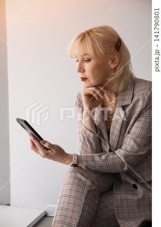 Senior blonde businesswoman in a plaid suit sits indoors, focused on her smartphone, thoughtfully browsing digital content and emails, conveying modern professional confidence Senior blonde businesswoman in a plaid suit sits indoors, focused on her smartphone, thoughtfully browsing digital content and emails, conveying modern professional confidence 141790801