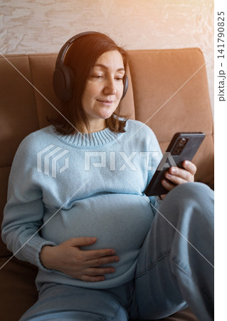 Pregnant woman wearing headphones while relaxing on the couch and listening to music at home Pregnant woman wearing headphones while relaxing on the couch and listening to music at home 141790825