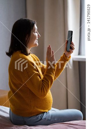 Pregnant woman sitting by a window using a smartphone in warm morning light Pregnant woman sitting by a window using a smartphone in warm morning light 141790829