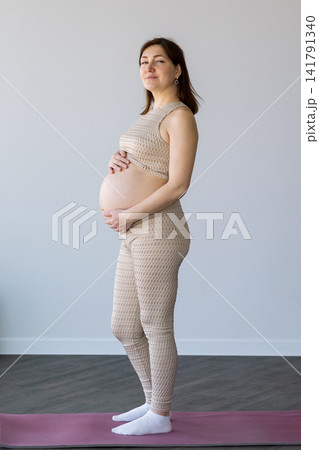 Pregnant woman standing on yoga mat touching belly Pregnant woman standing on yoga mat touching belly 141791340