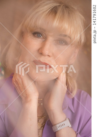 Mature blonde woman elegantly posing for a close up portrait, hands gently touching her neck, wearing a lavender blouse and layered gold necklaces, displaying a calm and thoughtful expression Mature blonde woman elegantly posing for a close up portrait, hands gently touching her neck, wearing a lavender blouse and layered gold necklaces, displaying a calm and thoughtful expression 141791682