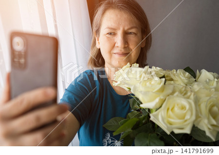 Mature woman happily capturing a selfie using her smartphone, holding a beautiful bouquet of white roses, celebrating special occasion or receiving a thoughtful gift 141791699