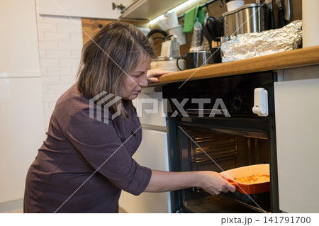 Woman prepares a homemade meal, carefully placing a dish with food into a hot oven, demonstrating domestic life and cooking activities in a cozy kitchen environment Woman prepares a homemade meal, carefully placing a dish with food into a hot oven, demonstrating domestic life and cooking activities in a cozy kitchen environment 141791700
