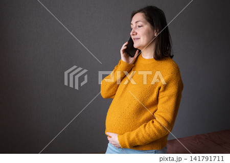 Pregnant woman in a mustard sweater holding her belly and talking on the phone Pregnant woman in a mustard sweater holding her belly and talking on the phone 141791711