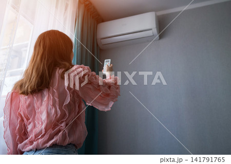 Woman adjusts split system air conditioner with remote control, cooling a sunny apartment for comfortable indoor climate and energy efficient relief during hot summer weather Woman adjusts split system air conditioner with remote control, cooling a sunny apartment for comfortable indoor climate and energy efficient relief during hot summer weather 141791765