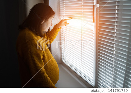 Pregnant woman standing by window blinds in warm morning light 141791789