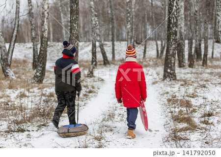 Two children walking in a snowy forest with sleds in winter clothing Two children walking in a snowy forest with sleds in winter clothing 141792050