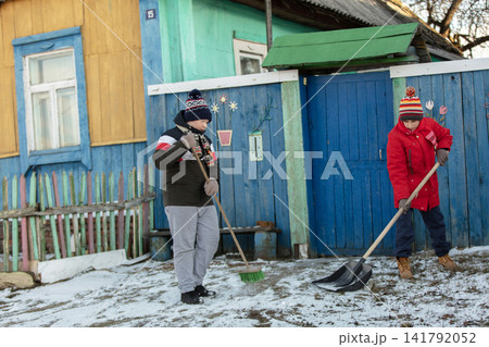 Two children shoveling snow in front of a colorful wooden house in winter attire 141792052
