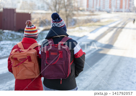 Schoolchildren with backpacks walk with their backs to the camera along a snowy road. Schoolchildren with backpacks walk with their backs to the camera along a snowy road. 141792057