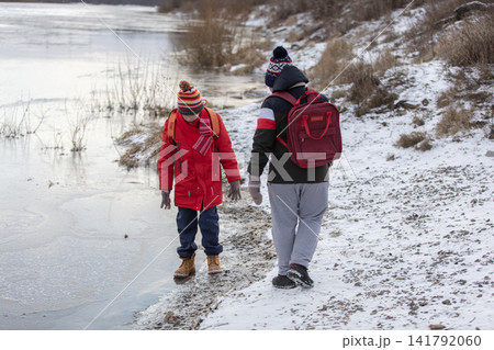 Two children explore the snowy river bank. One boy tries to walk on thin ice. Two children explore the snowy river bank. One boy tries to walk on thin ice. 141792060