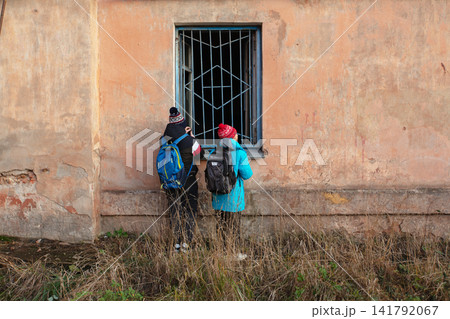Two people with backpacks and winter hats looking into a barred window of an old weathered building Two people with backpacks and winter hats looking into a barred window of an old weathered building 141792067