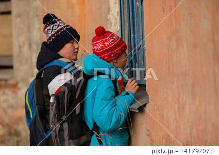 Two children in colorful winter jackets and hats look through the metal window bars of a dilapidated abandoned building 141792071