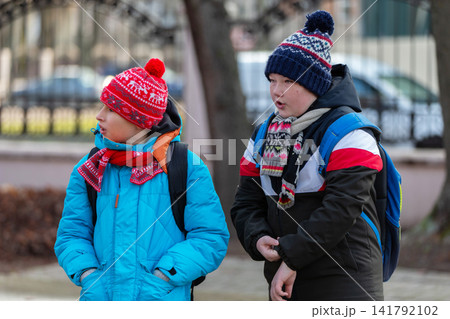 Children wear hats and jackets while standing outside on a cold day in a city park watching people pass by 141792102