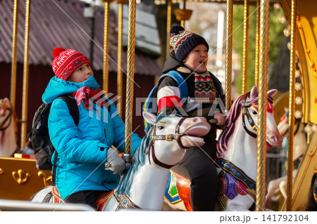 Kids enjoy a ride on a carousel at a fair in autumn while wearing warm clothing and hats Kids enjoy a ride on a carousel at a fair in autumn while wearing warm clothing and hats 141792104