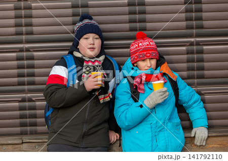 Two children drink warm beverages while leaning against a wall in a chilly outdoor setting during winter 141792110