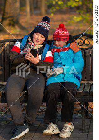 Two school age boys sitting on a bench and looking at a smartphone in the park 141792127