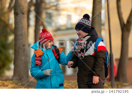 Two schoolboy boys with backpacks walk in an autumn park in bright jackets and hats, and leaves are falling around them. Two schoolboy boys with backpacks walk in an autumn park in bright jackets and hats, and leaves are falling around them. 141792195