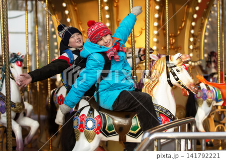 Schoolchildren with backpacks in winter clothes enjoy a ride on a carousel with colorful horses and bright lights Schoolchildren with backpacks in winter clothes enjoy a ride on a carousel with colorful horses and bright lights 141792221