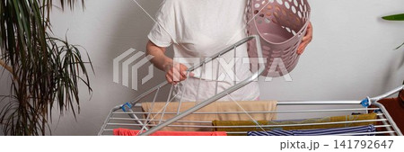 A 50-year-old woman hangs freshly washed laundry on a drying rack inside her room. She is holding a laundry basket filled with clothes, organizing her space, banner 141792647