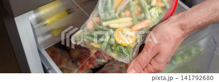 A man reaches into the refrigerator and takes out a bag of frozen vegetables. He is preparing to cook during the afternoon in a kitchen setting, banner A man reaches into the refrigerator and takes out a bag of frozen vegetables. He is preparing to cook during the afternoon in a kitchen setting, banner 141792680