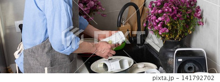 A man stands at the sink in his kitchen, cleaning dirty dishes and utensils. Flowers are placed on the counter, adding color to the scene in daylight, banner A man stands at the sink in his kitchen, cleaning dirty dishes and utensils. Flowers are placed on the counter, adding color to the scene in daylight, banner 141792702