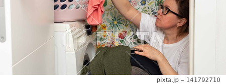 Woman stands in bathroom putting clothes into washing machine while surrounded by towels and laundry, carrying out daily household task with focus and care, banner Woman stands in bathroom putting clothes into washing machine while surrounded by towels and laundry, carrying out daily household task with focus and care, banner 141792710