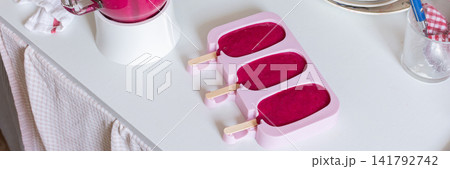 In a kitchen, a blender is mixing strawberries, bananas, and beets. Ice cream molds sit on the table filled with the mixture. A ripe banana and a beet are nearby, banner 141792742