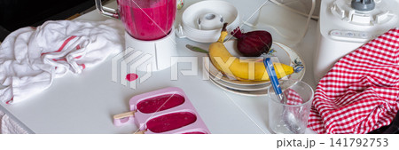 In a kitchen, a blender is mixing strawberries, bananas, and beets. Ice cream molds sit on the table filled with the mixture. A ripe banana and a beet are nearby, banner 141792753