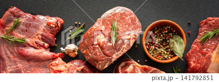 Several types of beef steak are laid out on a dark surface with fresh herbs and spices. Ingredients include garlic, pepper, and oil, showing preparation for cooking in a kitchen, banner 141792851