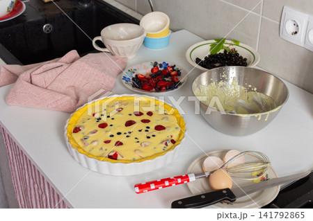 A person prepares a pie filled with strawberries, currants, and blueberries. The pie crust is filled with a creamy mixture. Ingredients are placed around the pie on a kitchen counter A person prepares a pie filled with strawberries, currants, and blueberries. The pie crust is filled with a creamy mixture. Ingredients are placed around the pie on a kitchen counter 141792865