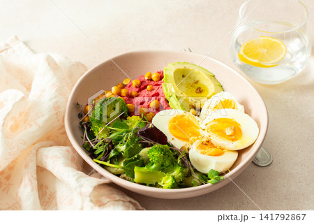 Bowls of beet hummus contain chickpeas, boiled eggs, fresh broccoli, and microgreens salad. Glasses of water with lemon slices add freshness to the table 141792867