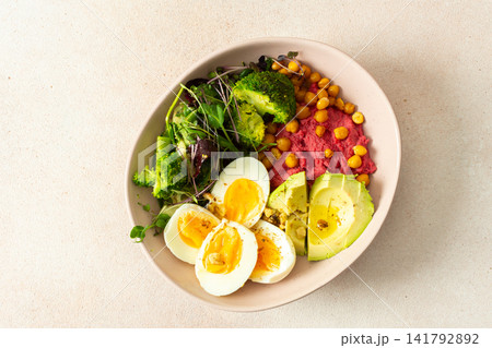 A bowl holds beet hummus topped with chickpeas, fresh broccoli, and microgreens salad alongside slices of avocado and boiled eggs. This meal is nutritious and filling 141792892
