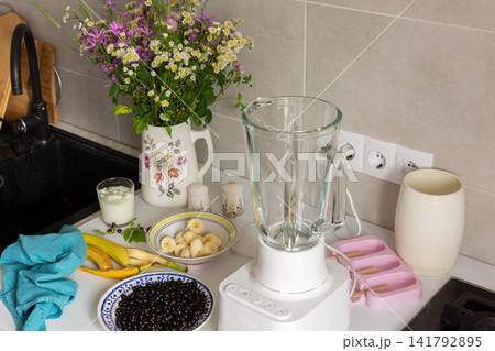 In a kitchen, bananas and currants are placed around a blender. There are bowls and containers ready for making homemade ice cream with fresh ingredients 141792895