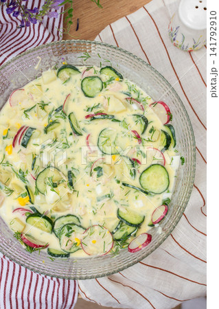 A bowl of okroshka filled with potatoes, chicken breast, cucumbers, herbs, and boiled eggs sits on a table. Nearby, a floral vase adds color to the setting 141792910