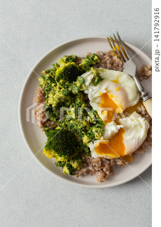A healthy breakfast bowl features green buckwheat topped with pesto, steamed broccoli, and poached eggs. A fork rests beside the dish ready for a nutritious meal A healthy breakfast bowl features green buckwheat topped with pesto, steamed broccoli, and poached eggs. A fork rests beside the dish ready for a nutritious meal 141792916