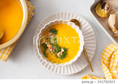 Pumpkin cream soup sits in a bowl with toppings next to a dish of baked hake and lemon. A spoon rests beside the soup in a clean dining area Pumpkin cream soup sits in a bowl with toppings next to a dish of baked hake and lemon. A spoon rests beside the soup in a clean dining area 141792923