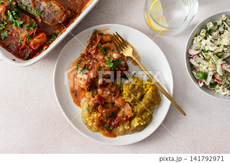 Plates hold amaranth, millet, and quinoa porridge served with baked white hake in tomato sauce. Fresh salad is beside the porridge. A bowl of kiwi sits nearby for dessert 141792971