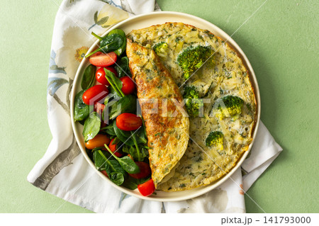 A potato omelette made with eggs and mixed greens is served on a white plate. Spinach, cherry tomatoes, and broccoli are arranged on the side for a healthy breakfast option 141793000