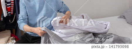 A man aged 53 is in his bedroom folding and changing bed linens. He sits on the bed with freshly cleaned sheets and a duvet beside him, focused on the task at hand, banner A man aged 53 is in his bedroom folding and changing bed linens. He sits on the bed with freshly cleaned sheets and a duvet beside him, focused on the task at hand, banner 141793098